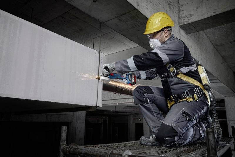 A construction worker on scaffolding is cutting a concrete beam with an electric saw. He is wearing protective clothing and a hard hat.