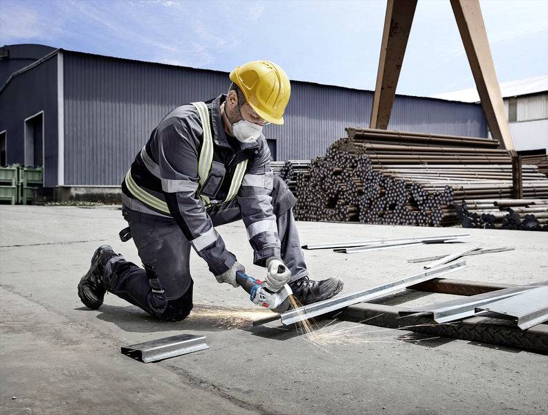 A construction worker wearing a hard hat and safety mask is cutting metal with an angle grinder on a building site. Stacks of metal beams are visible in the background.