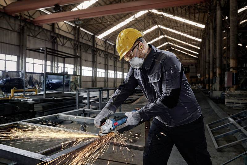 A worker in protective clothing grinds metal with an angle grinder in a large factory hall, sparks flying.