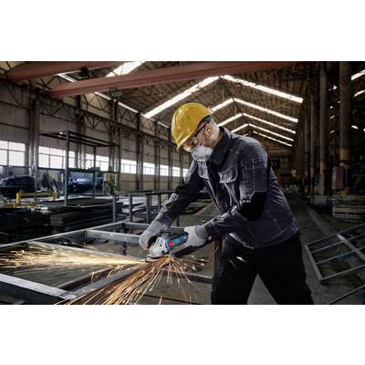 A worker in protective clothing grinds metal with an angle grinder in a large factory hall. Sparks are flying from the workpiece.