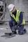 A worker is kneeling on a building site, wearing a hard hat and high-visibility vest, and using an electrical device to secure a screw.