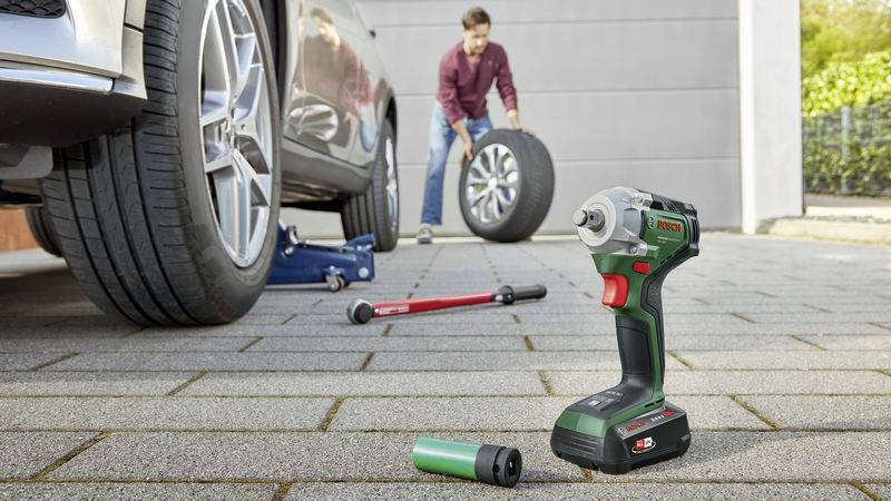 A man is changing a car tyre in a driveway, with an electric power drill lying on the ground in front of him.