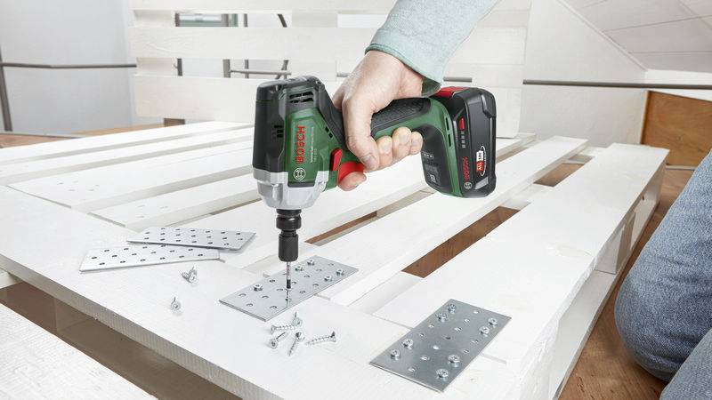 A person is operating a green cordless drill, fastening metal plates with screws onto a white wooden pallet.