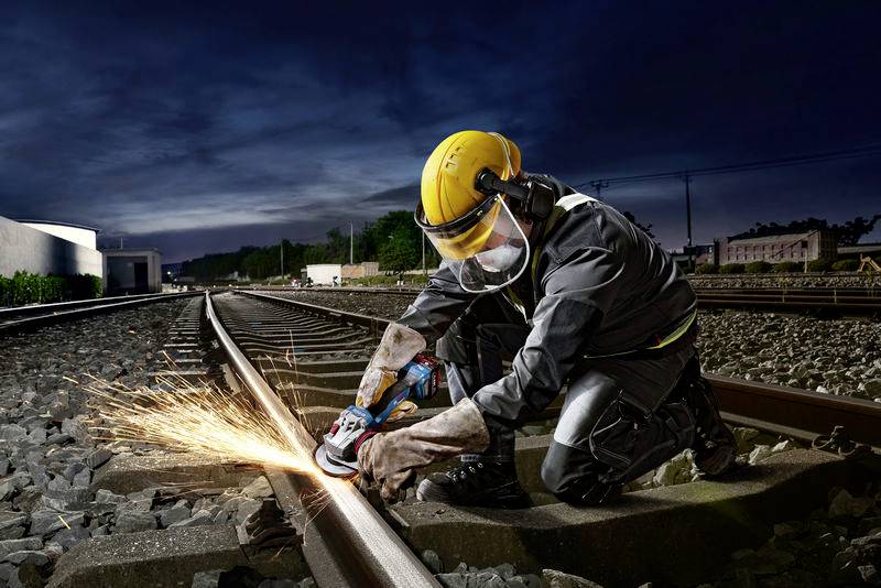 A worker in protective clothing and a hard hat is grinding a rail along a track at night. Sparks are flying from the grinding machine.