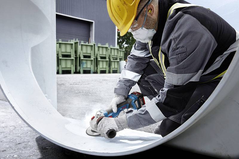 A worker in protective clothing is grinding the inner surface of a large pipe on a construction site. Green containers are visible in the background.