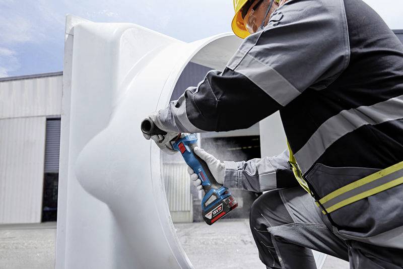 A worker wearing a hard hat is using an electric tool to work on a large white pipe. A building can be seen in the background.