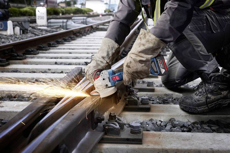 'Worker grinds railway tracks with an angle grinder, sparks flying. Worker is wearing safety clothing.'