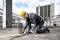 Worker on a building site, kneeling and cutting metal rods with an angle grinder. Scaffolding and unfinished building in the background.