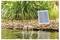A small solar panel powers a fountain in a pond, surrounded by rocks and tall grasses. The fountain creates a gentle water spray.