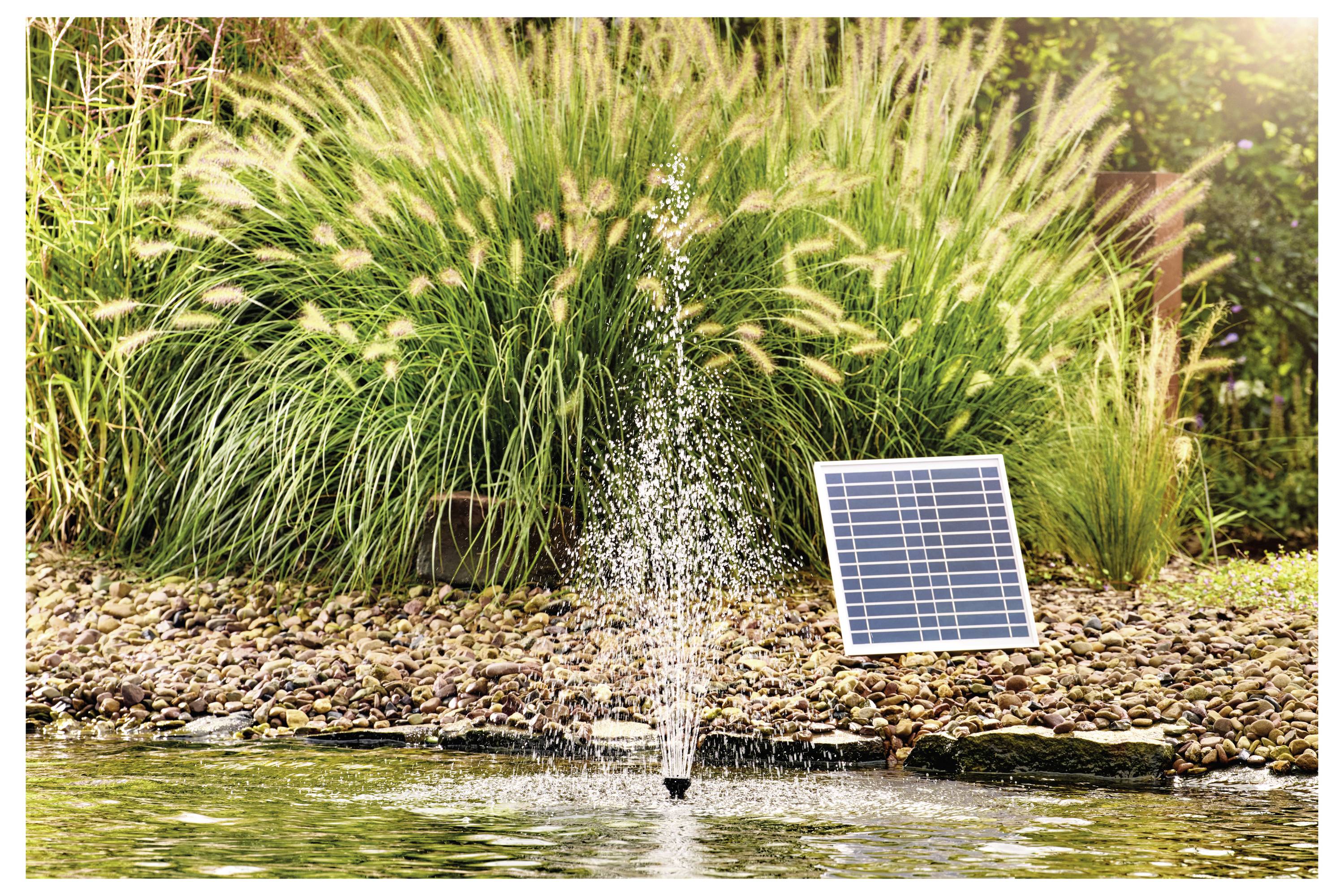 A small pond fountain surrounded by rocks, powered by a solar panel placed on the ground beside it, with tall grass in the background.