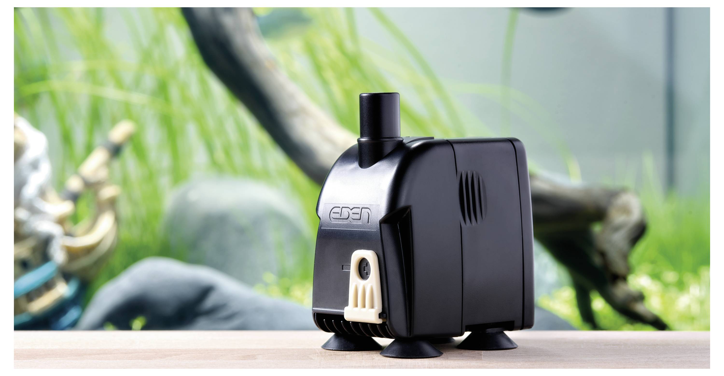 A small black aquarium pump with a nozzle sits on a light wooden surface. Blurred aquarium plants and rocks are in the background.