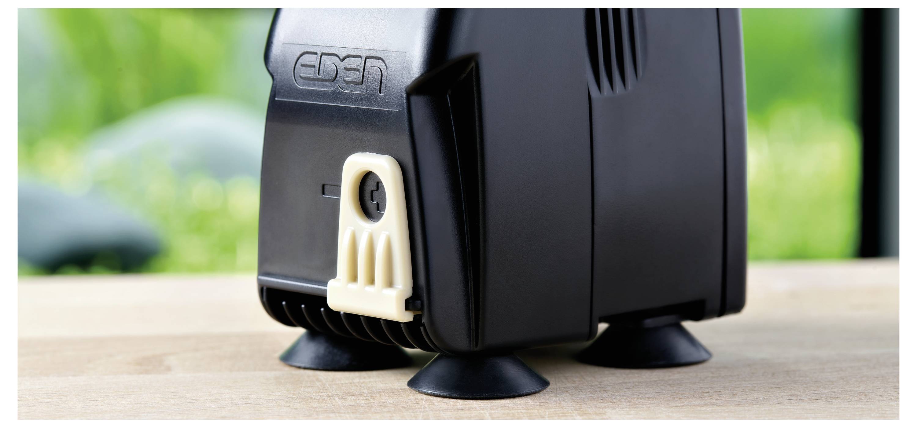 Close-up of a black machine with a beige plug cover, supported by suction cups on a wooden surface. Green blurred background.