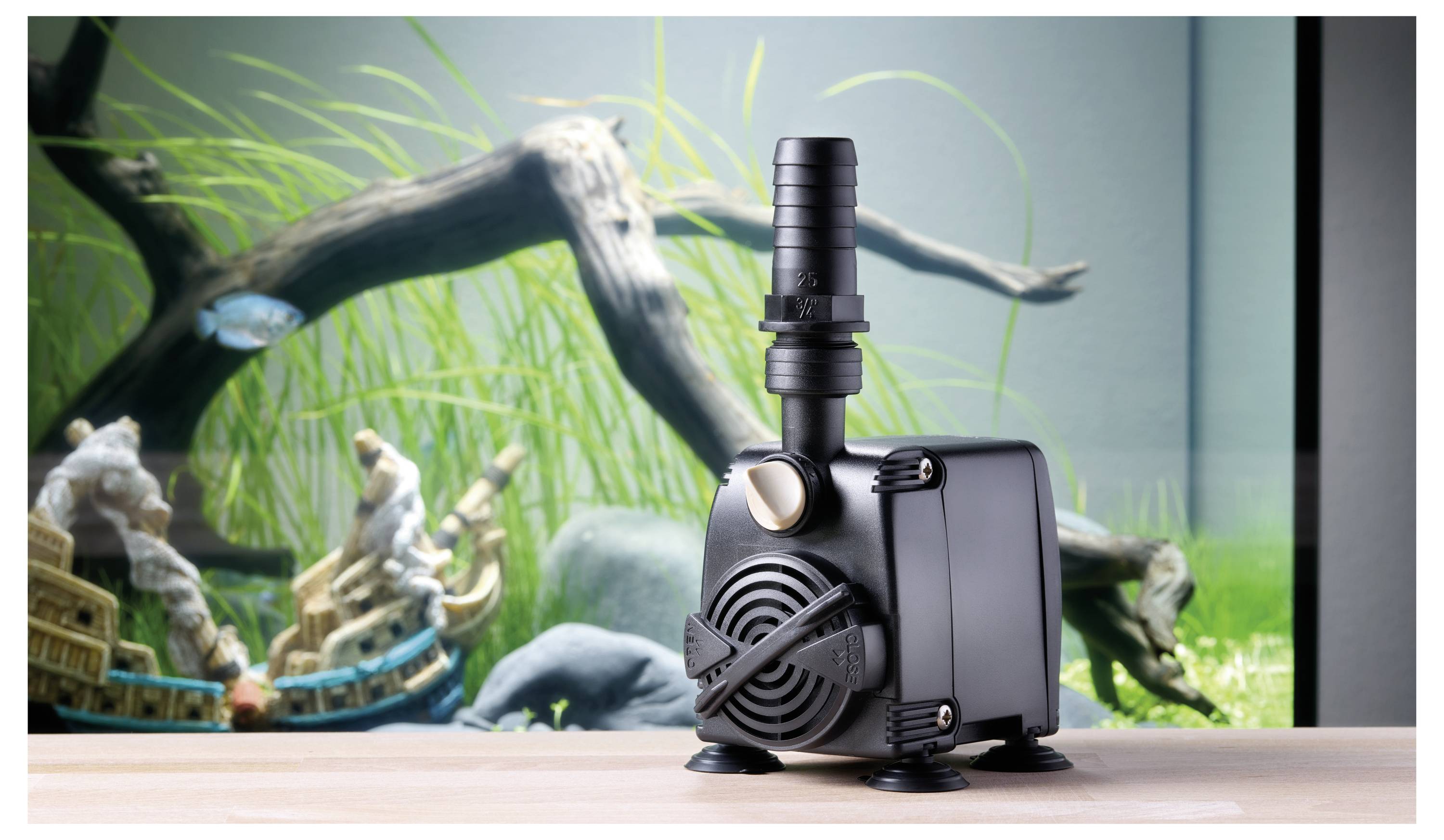 A black water pump is prominently displayed on a tabletop, with a blurry aquarium background showing green plants and a decorative arch.