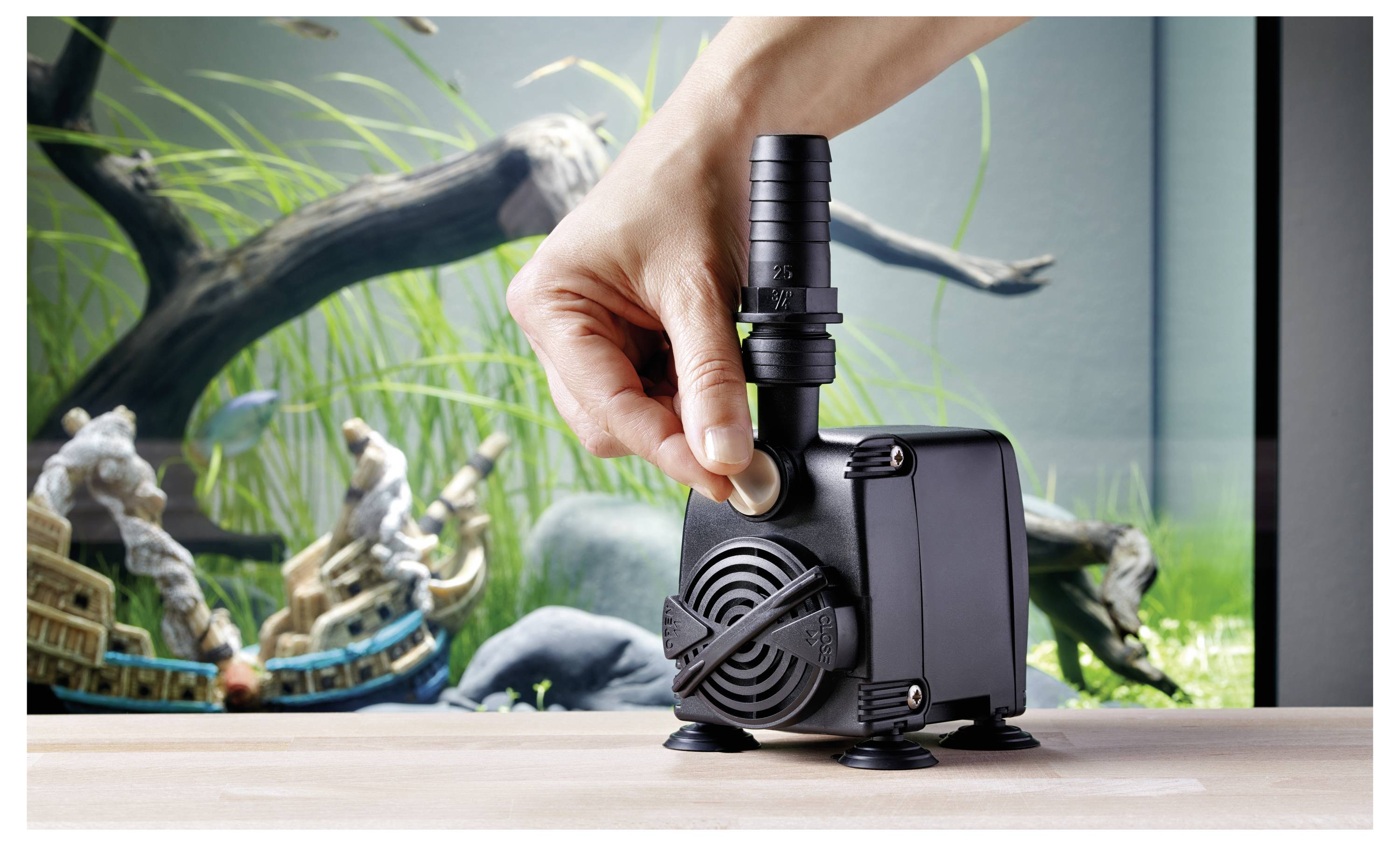 A hand adjusts a knob on a black aquarium pump, placed on a table with a fish tank in the background, showcasing aquatic plants and ornaments.