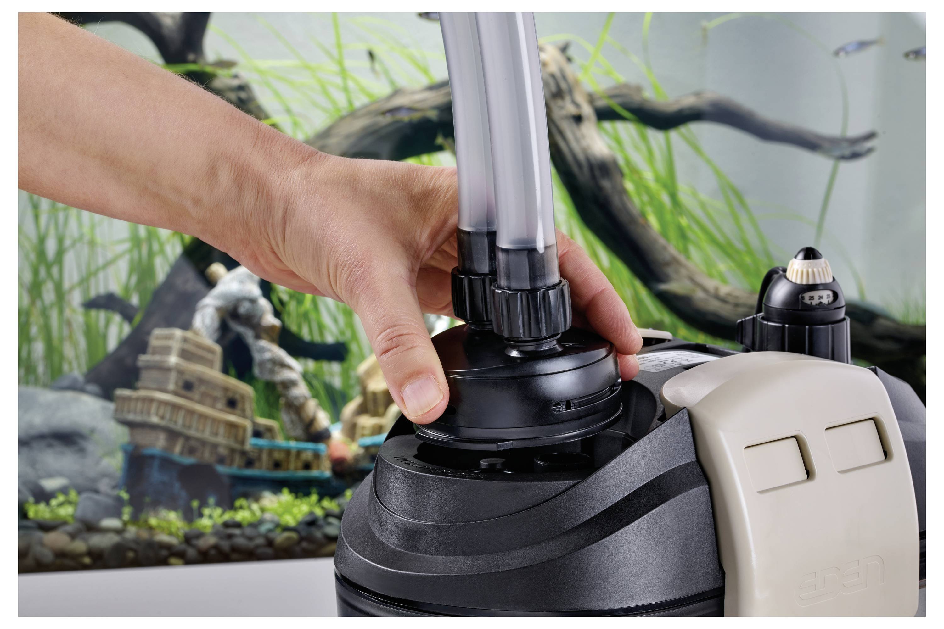 A hand assembles a filter unit connected to tubes in front of an aquarium with plants and decorative structures.