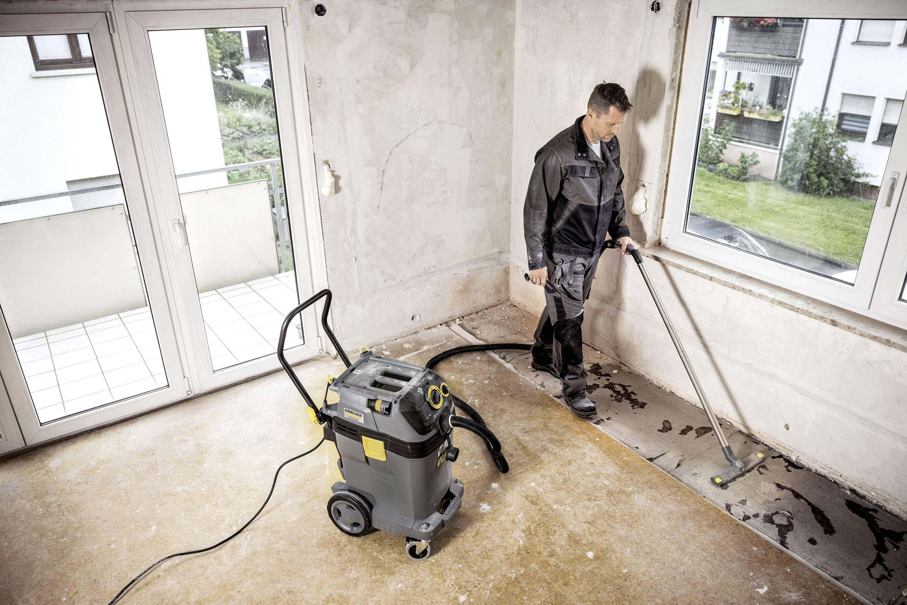 A man is cleaning an empty room with a wet vacuum cleaner. The floor is partially damp, and the window offers a view of the garden.