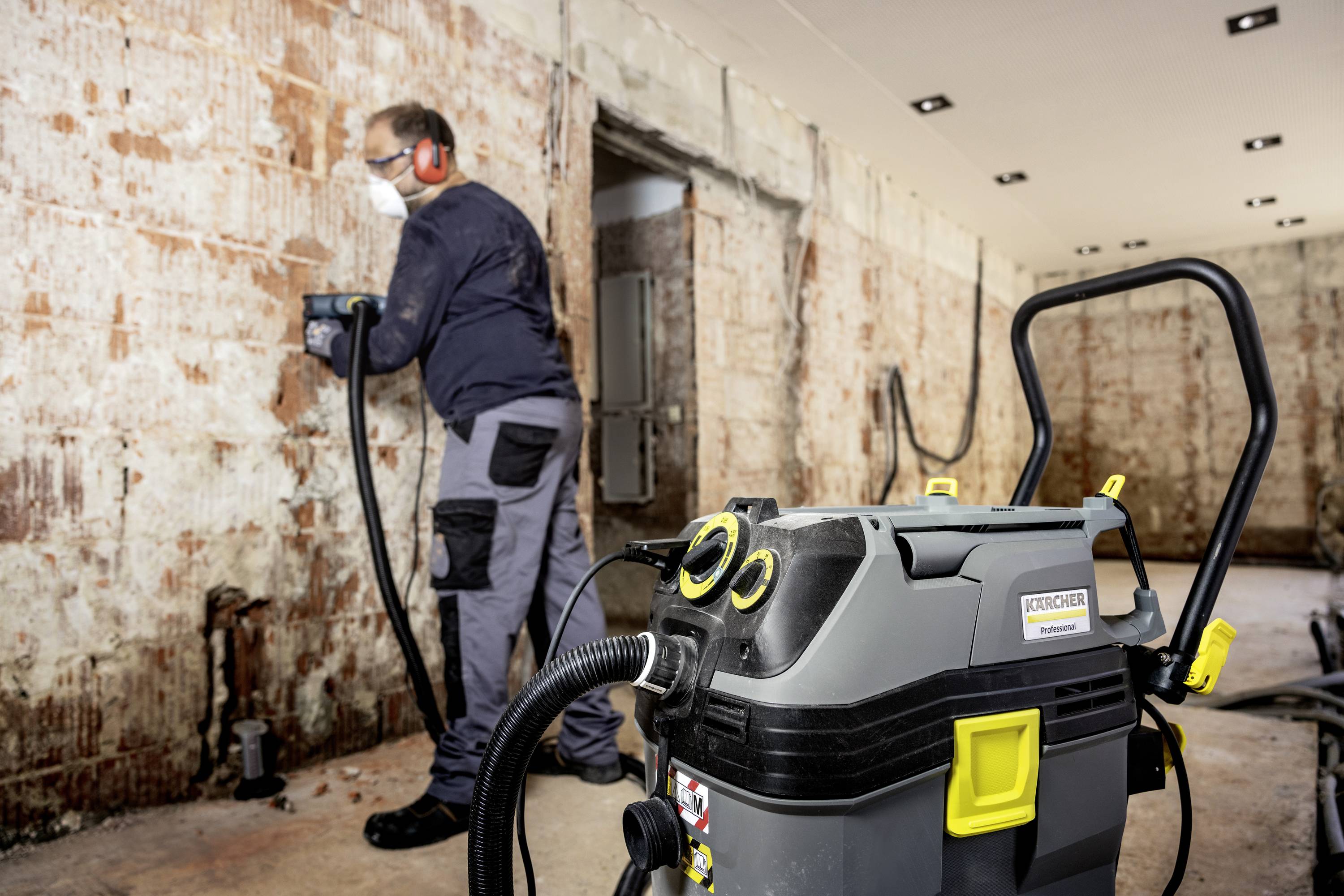 A tradesman is removing old plaster from a wall using a drill hammer. In the foreground, an industrial vacuum cleaner is sucking up dust.