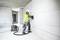 A construction worker in protective clothing is using an electrical tool in a partially renovated room. A vacuum unit is connected.