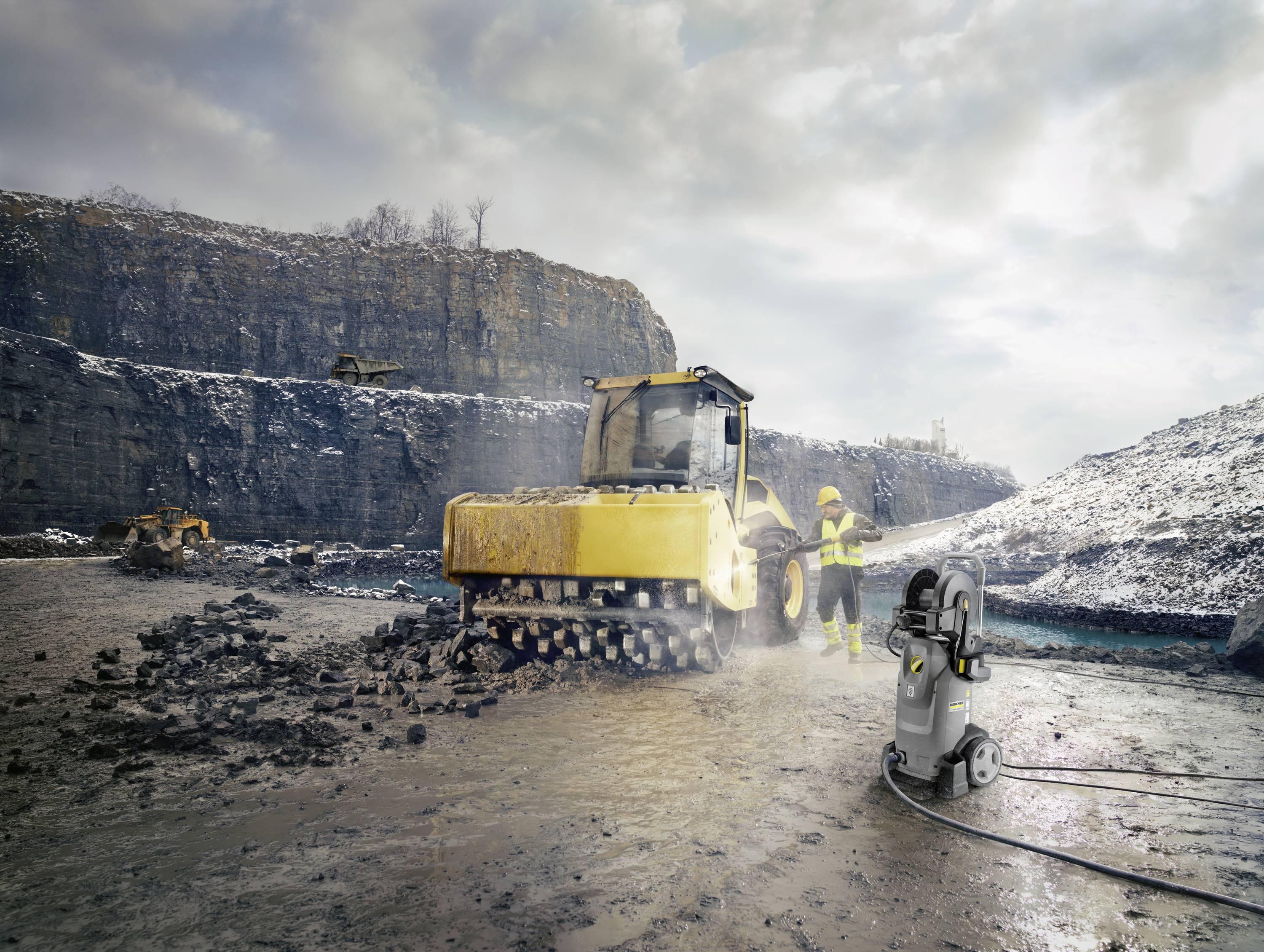 Two construction workers in protective clothing are operating a yellow road roller compactor on a rocky quarry site under cloudy skies.