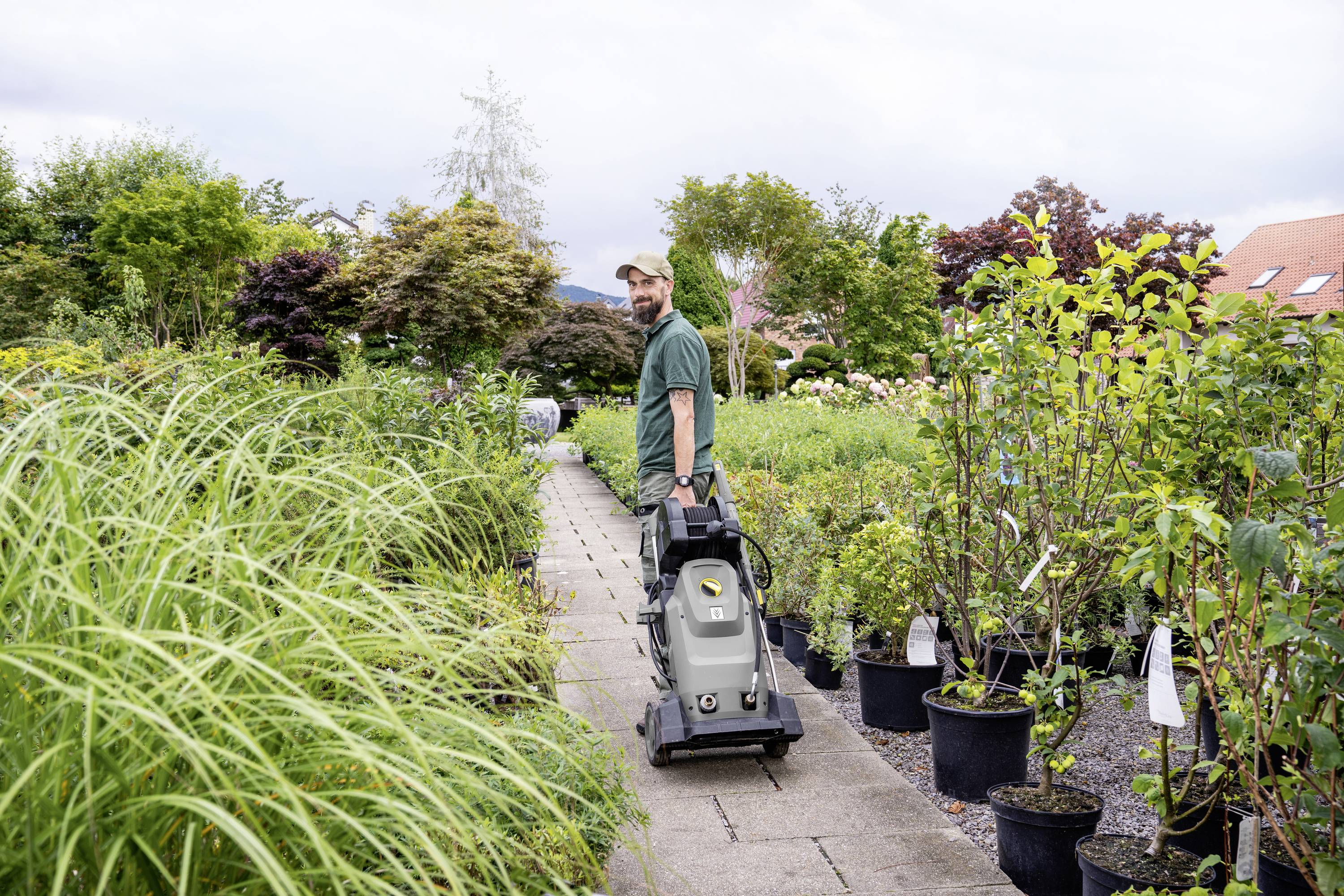 A man is pulling a lawnmower along a narrow path in a garden or plant centre. Numerous potted plants surround him.