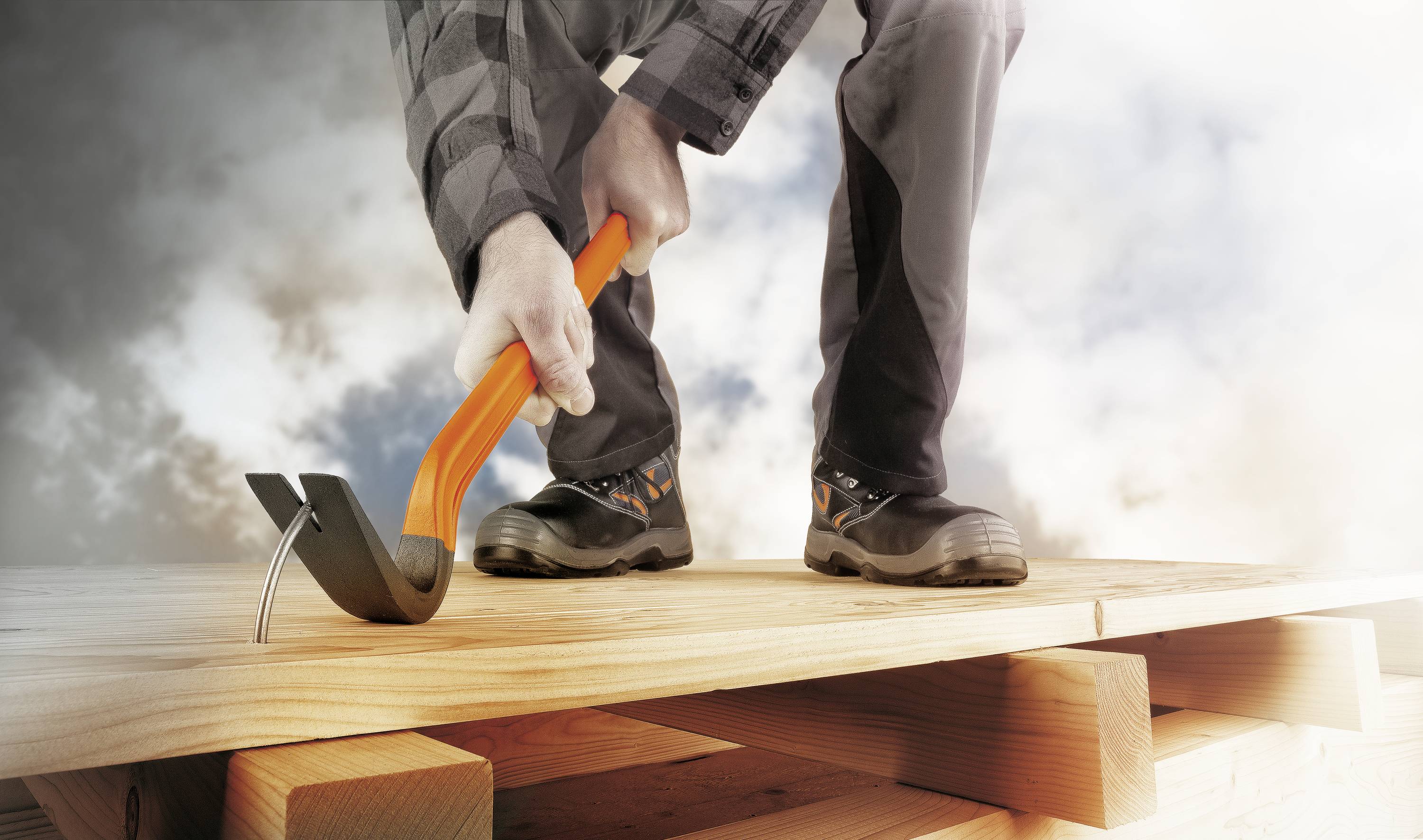A person is removing a board from a woodpile using a crowbar. They are wearing work clothes and sturdy shoes.