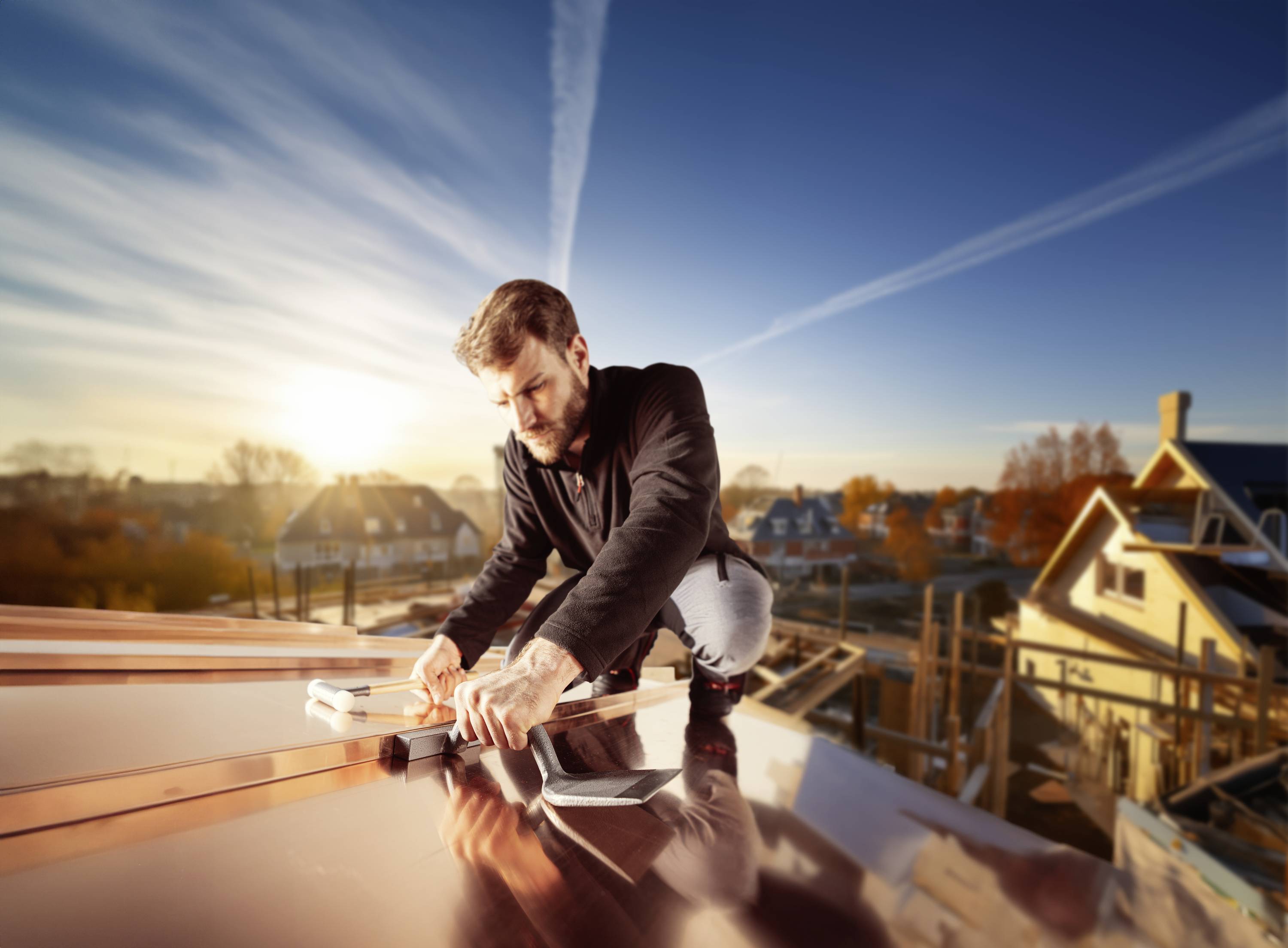 A man is working on the roof of a house at sunset. He is using tools to secure the roof surface.