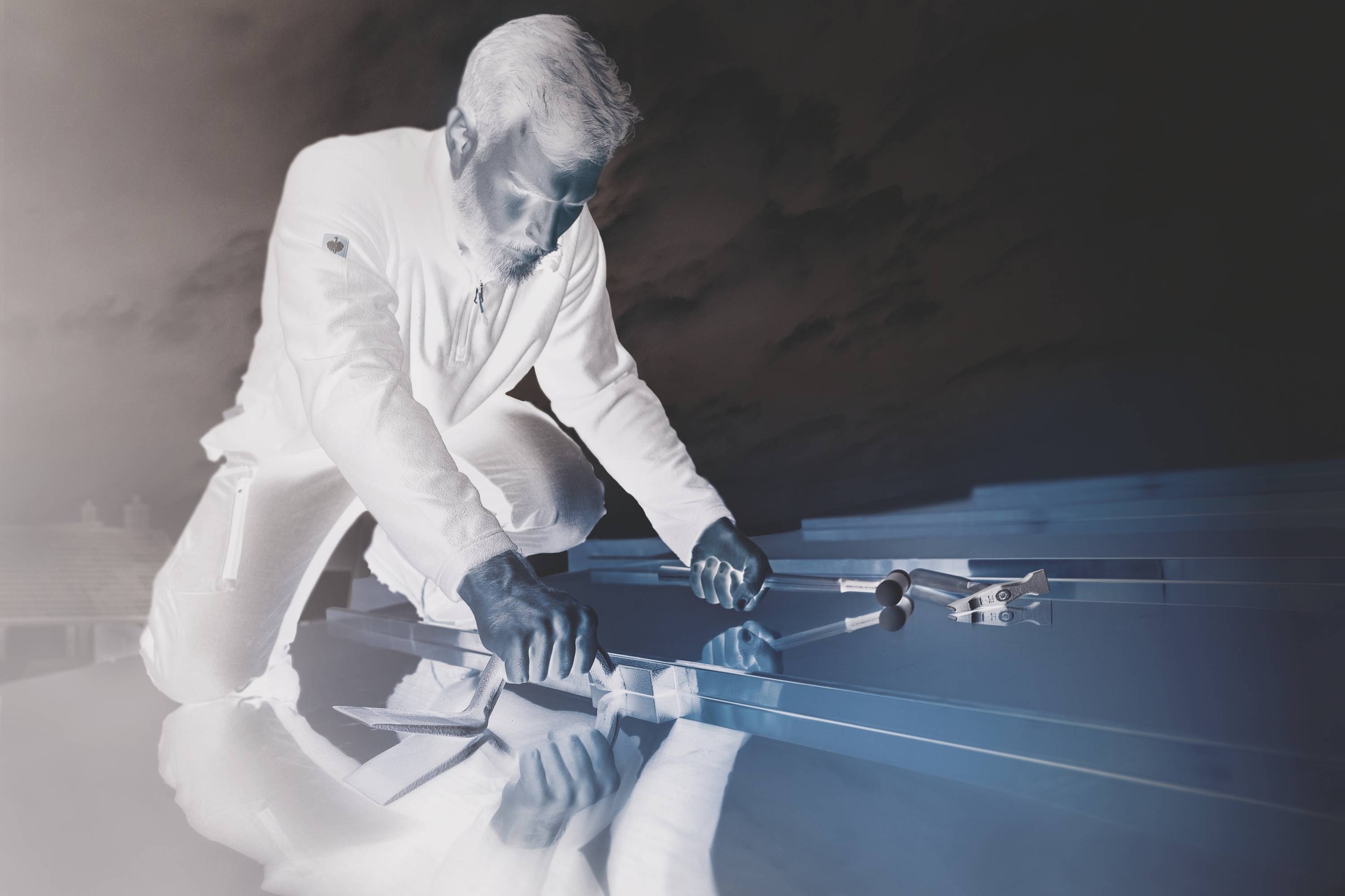 A tradesman kneels on a roof and works with tools on solar panels, beneath a cloudy sky.