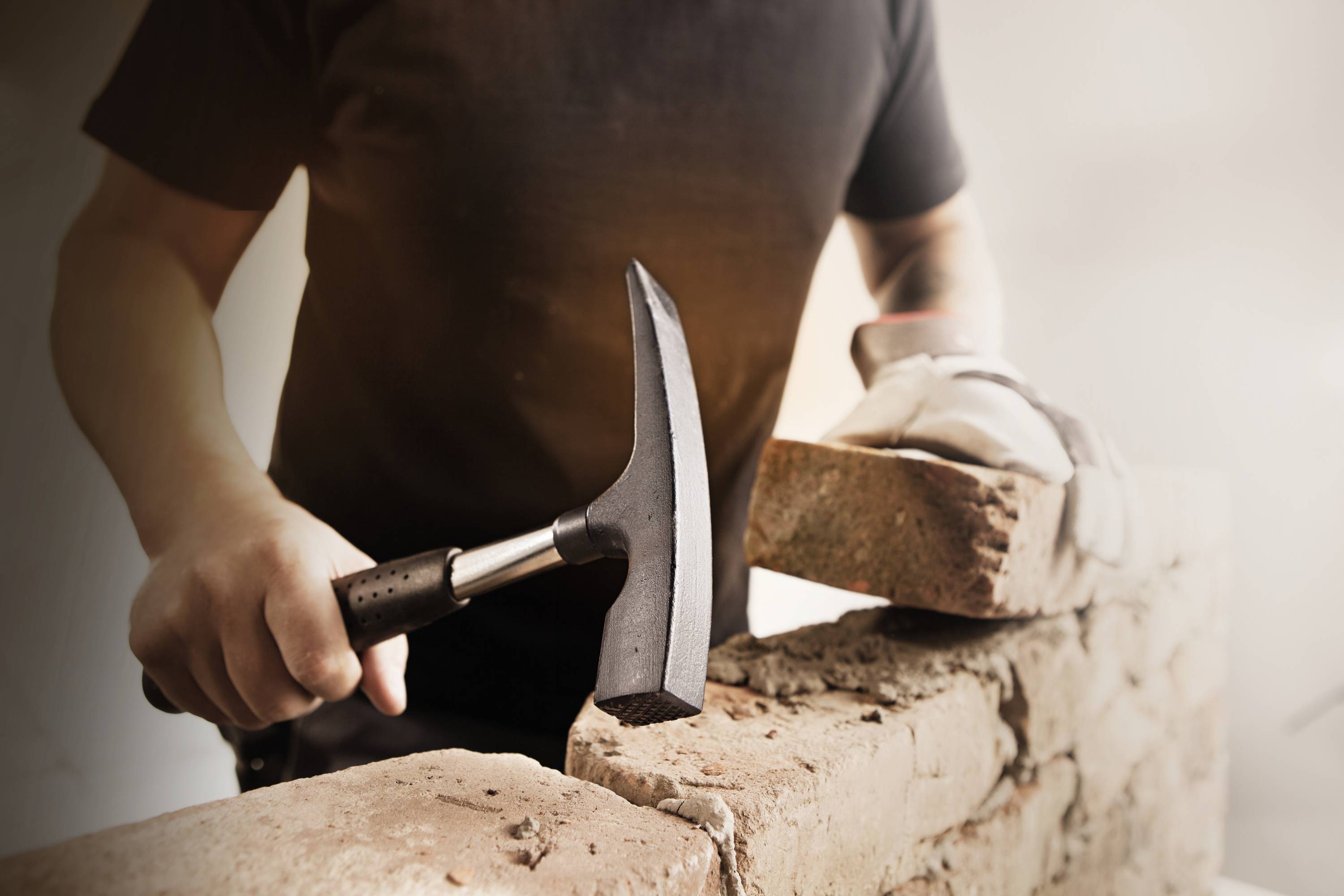A person who builds brickwork; holding a hammer in one hand and a brick in the other, under oblique lighting.