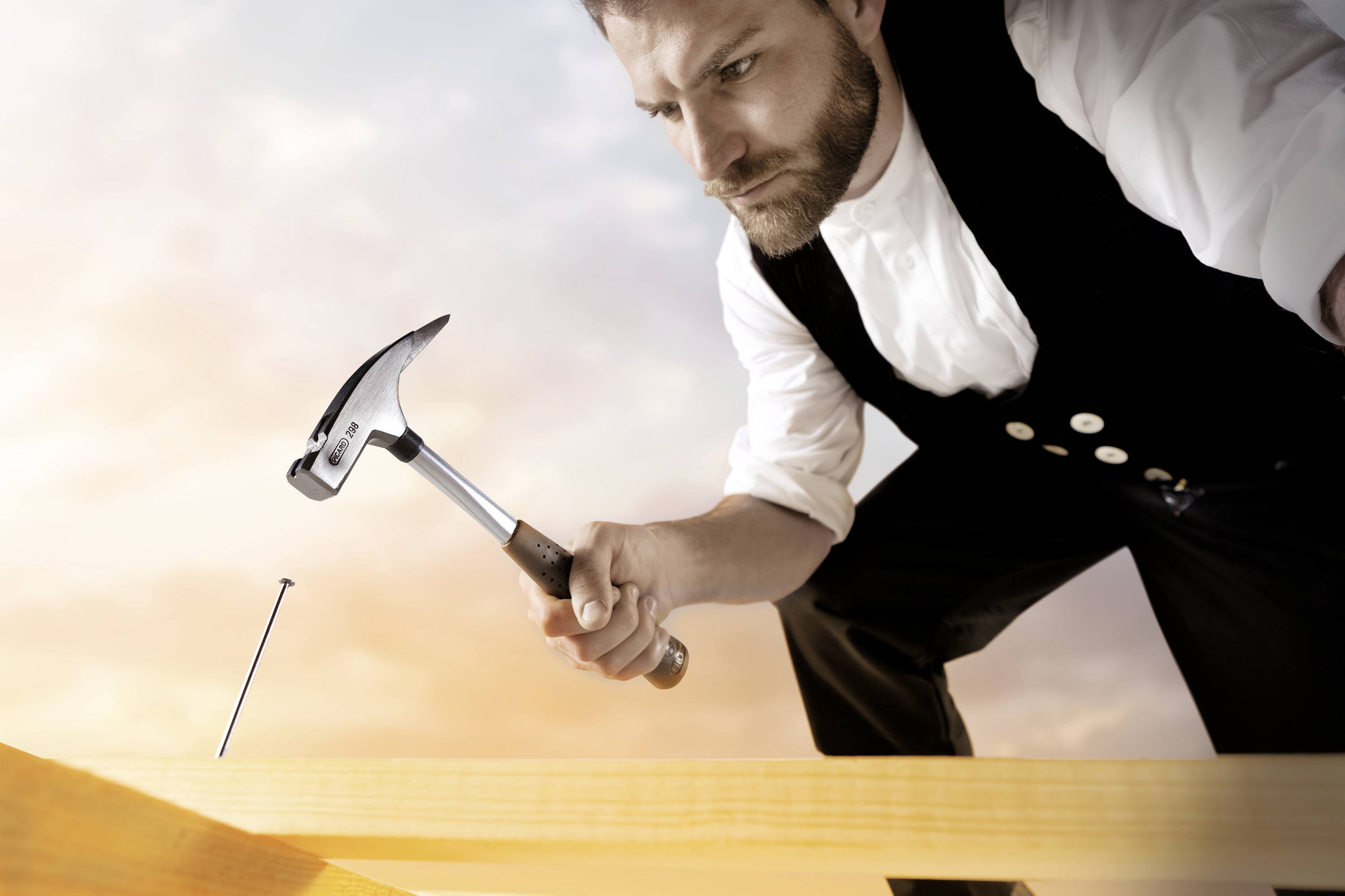 A man in traditional craftsman's attire hammers a nail into a wooden board. He is intensely focused on his work.
