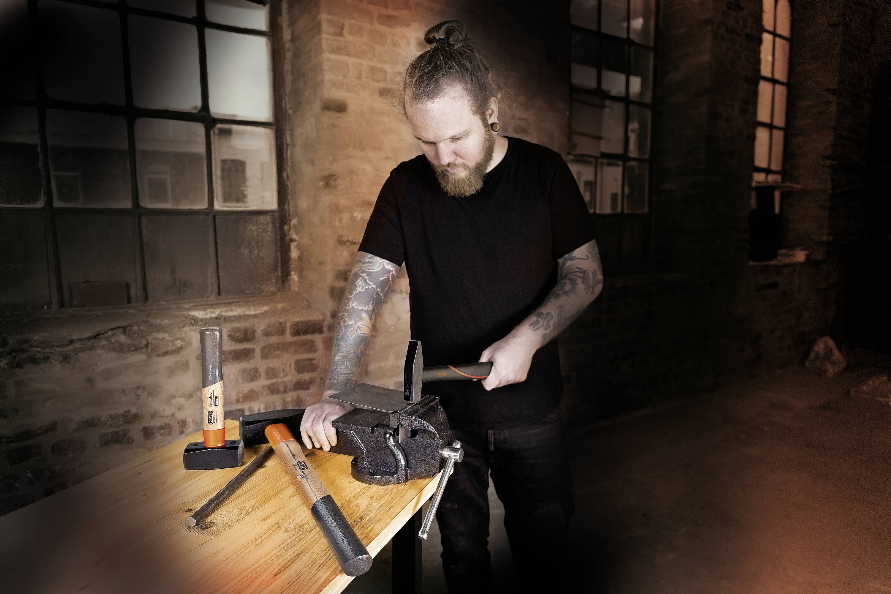 A person with tattoos is working in a workshop. They are using a file on a vice attached to a wooden workbench.