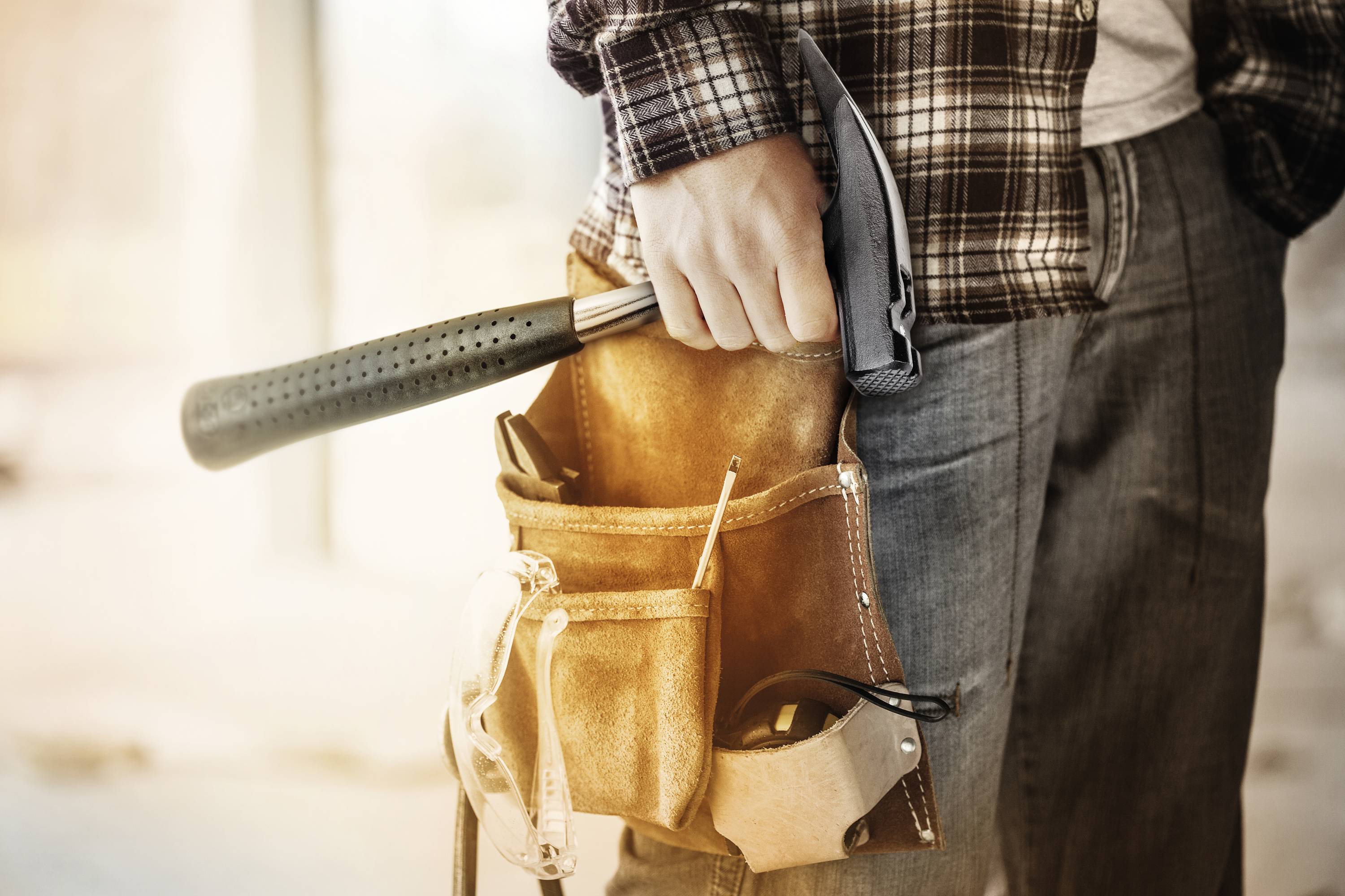 A person in workwear is holding a hammer and wearing a tool belt.