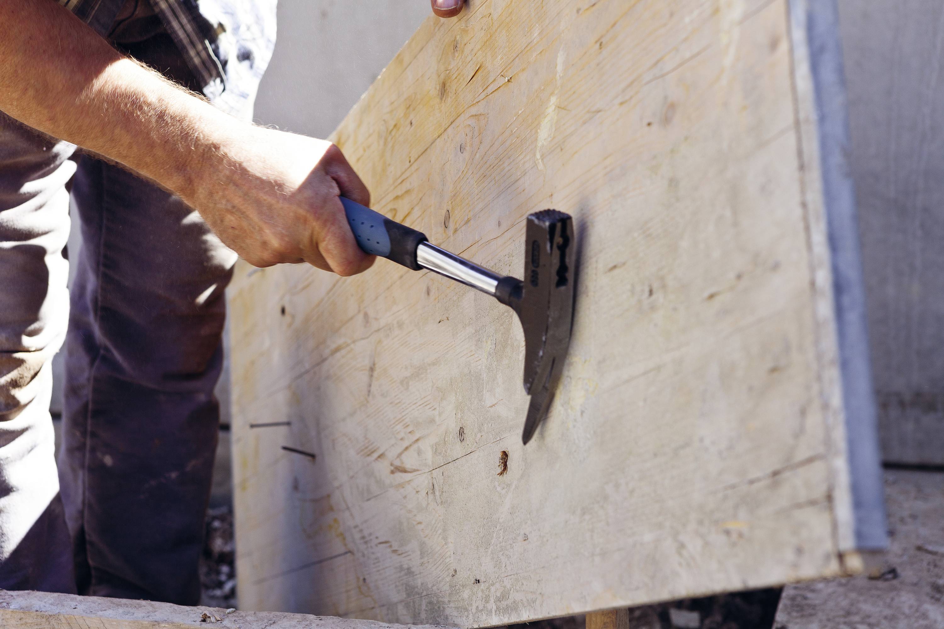 A person is using a hammer to drive a nail into a wooden board. The scene depicts craftsmanship outdoors.