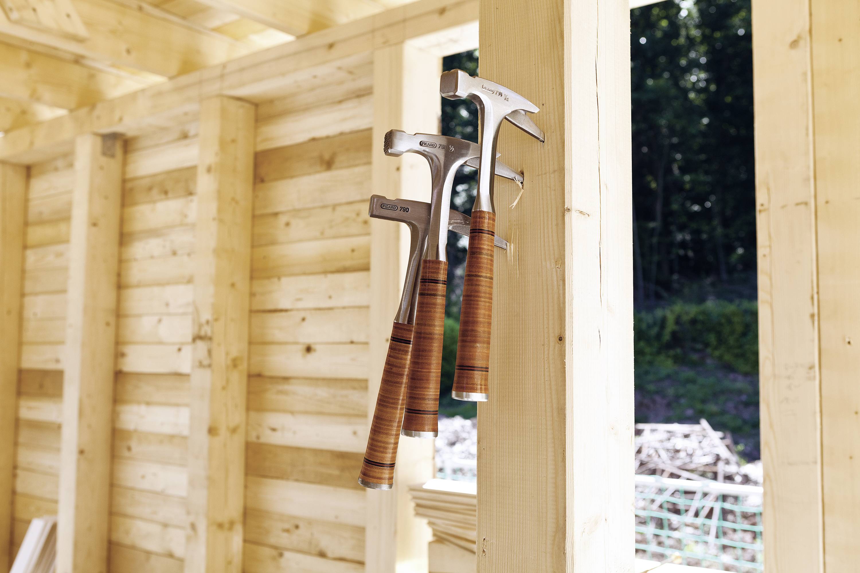 Three hammers with wooden handles hang side by side on a wooden wall in a building under construction, surrounded by natural light.