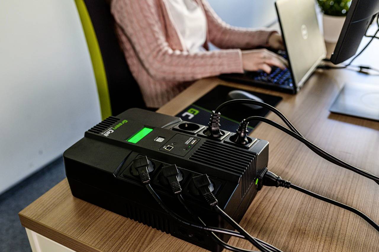 A person is working on a laptop next to an uninterruptible power supply (UPS) on a desk. Cables are connected.