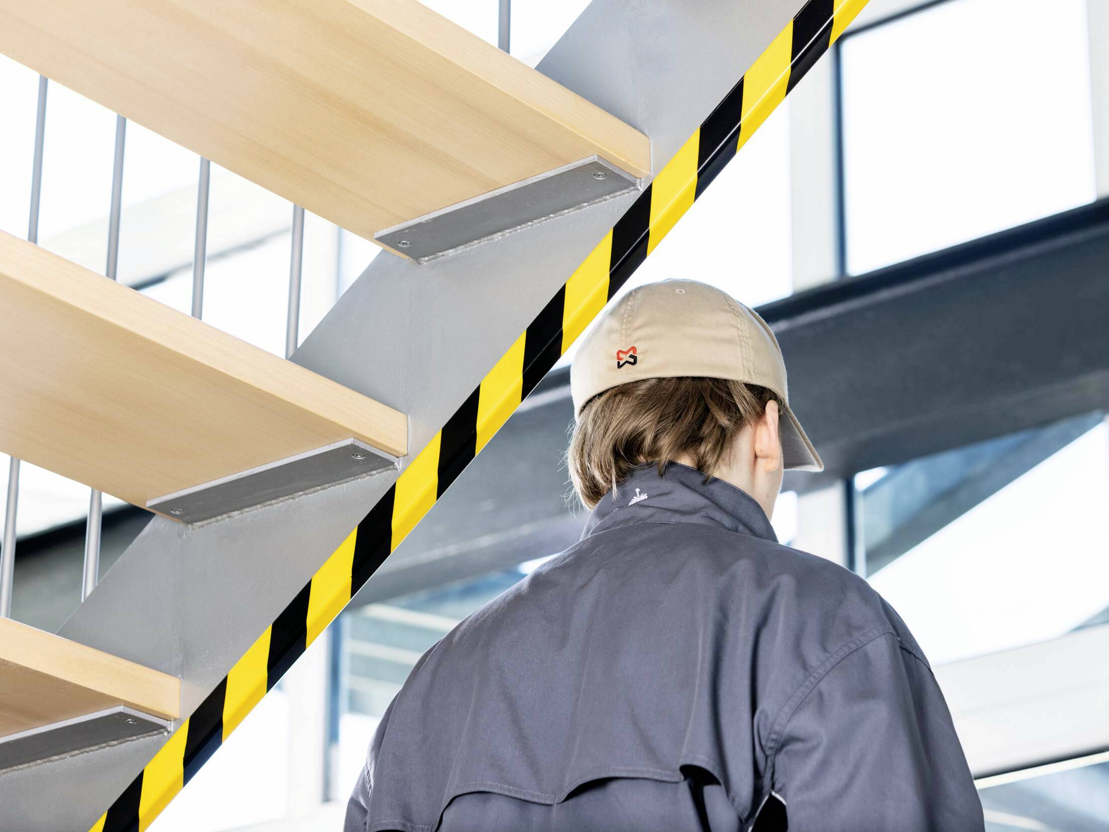 'Person in work attire standing in front of a staircase with yellow and black warning stripes. Modern architecture in the background.'