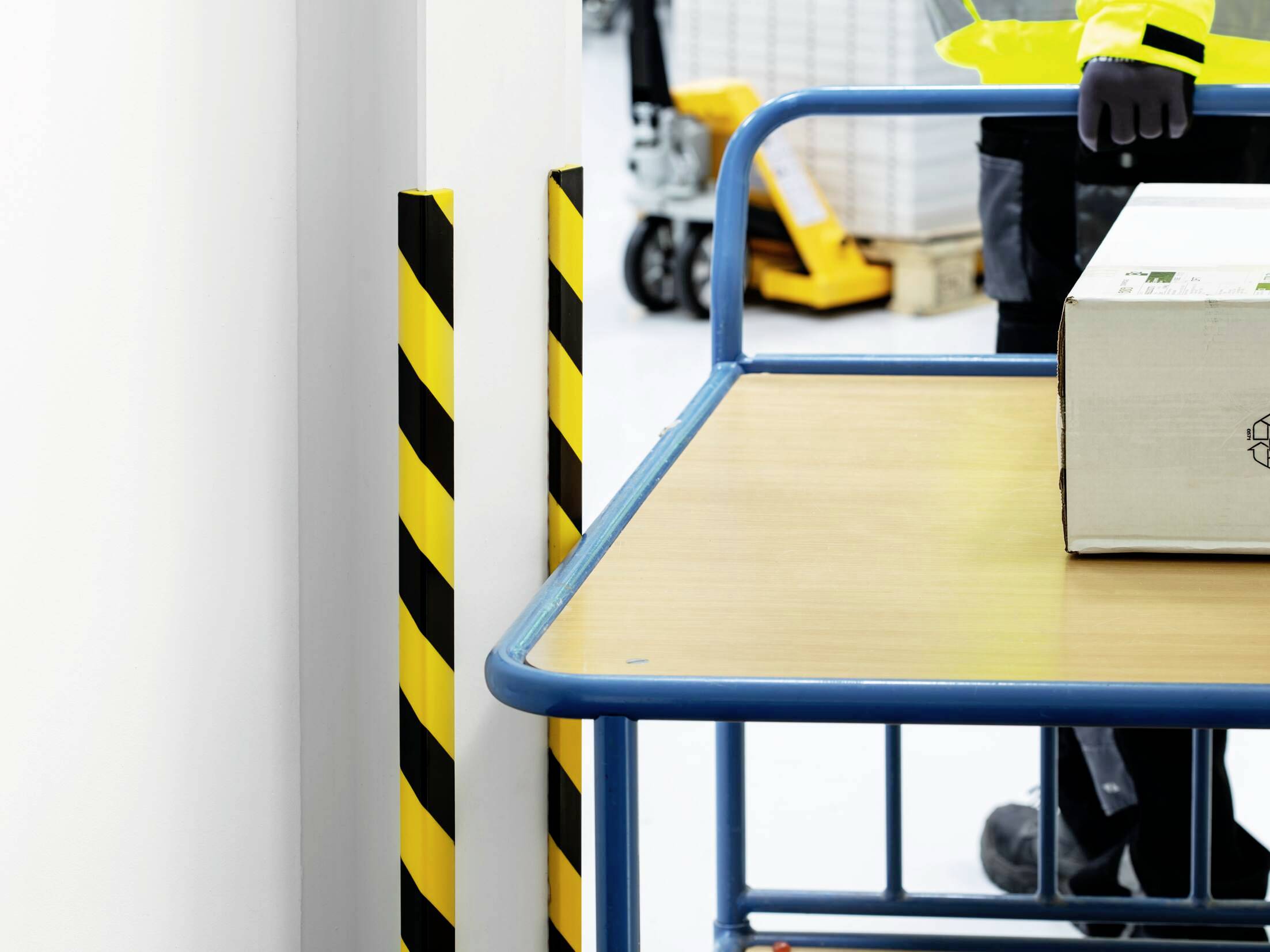 A worker is pushing a blue trolley with a cardboard box on top. In the background, yellow frames and part of a forklift are visible.