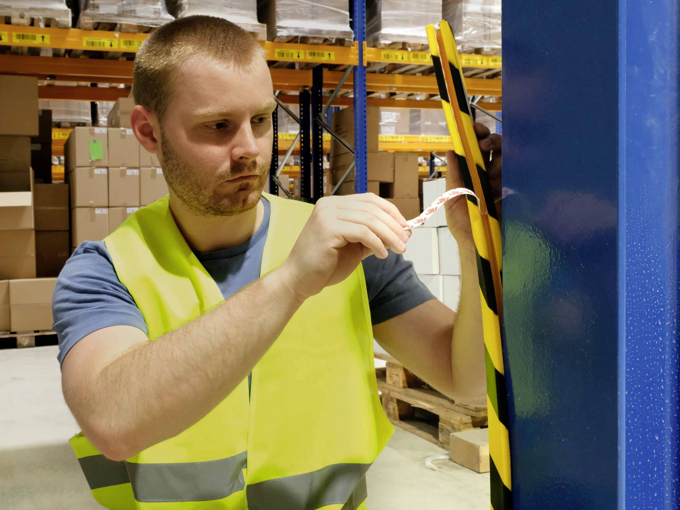 A man wearing a high-visibility vest is securing a tape to a pillar in a warehouse to implement a safety measure.
