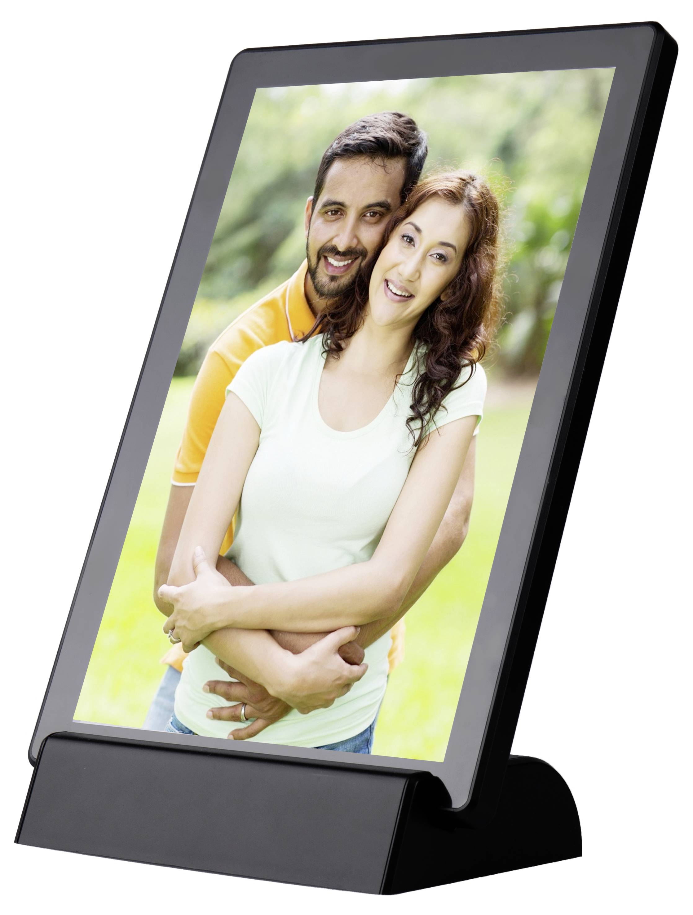 A smiling couple stands outdoors. He embraces her from behind, both looking happy and relaxed. The background is green and blurred.