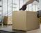 A person is opening a large brown cardboard box with a black and red utility knife in a warehouse with shelves.