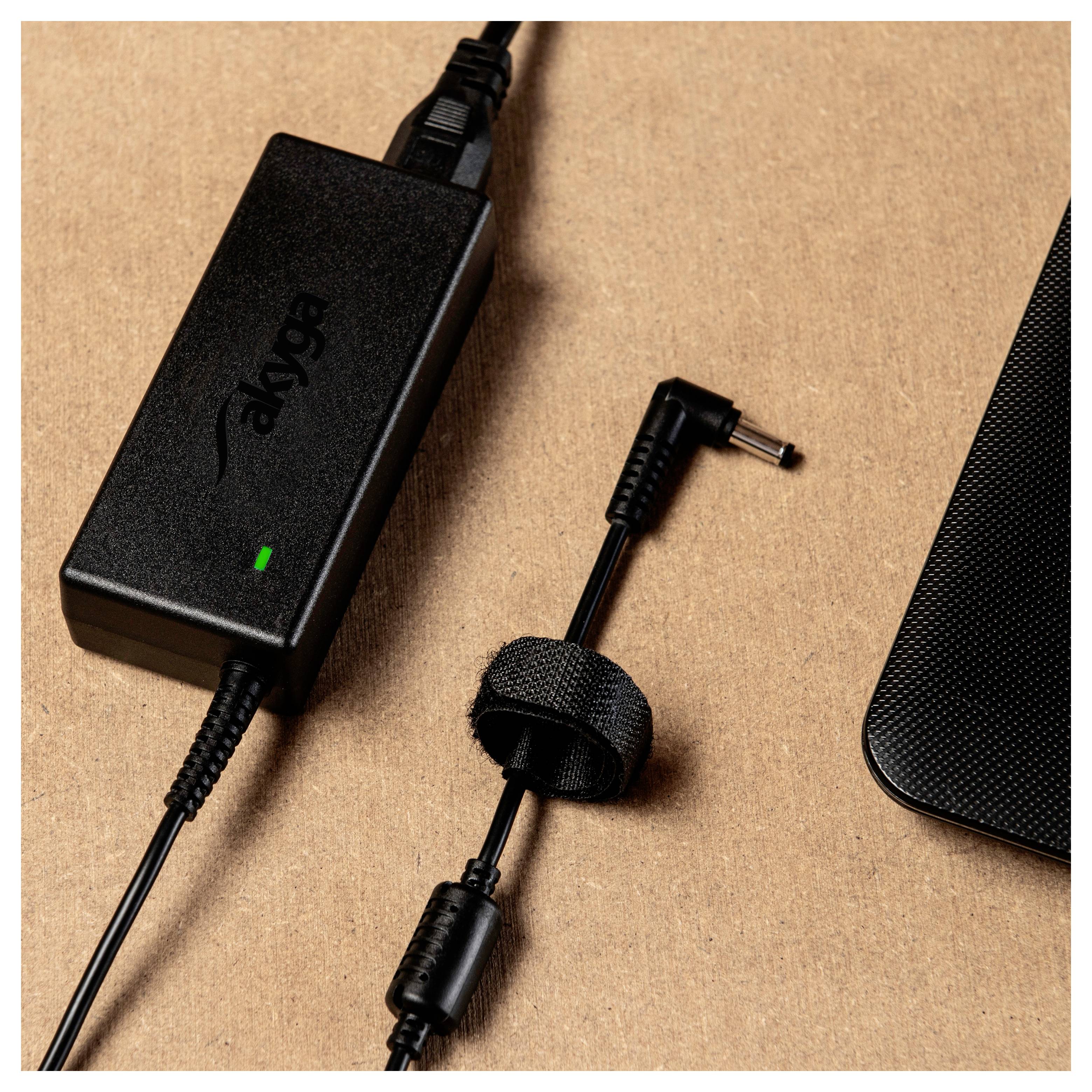 A black power supply with cable and plug lies on a brown surface. A strap holds the cable together.