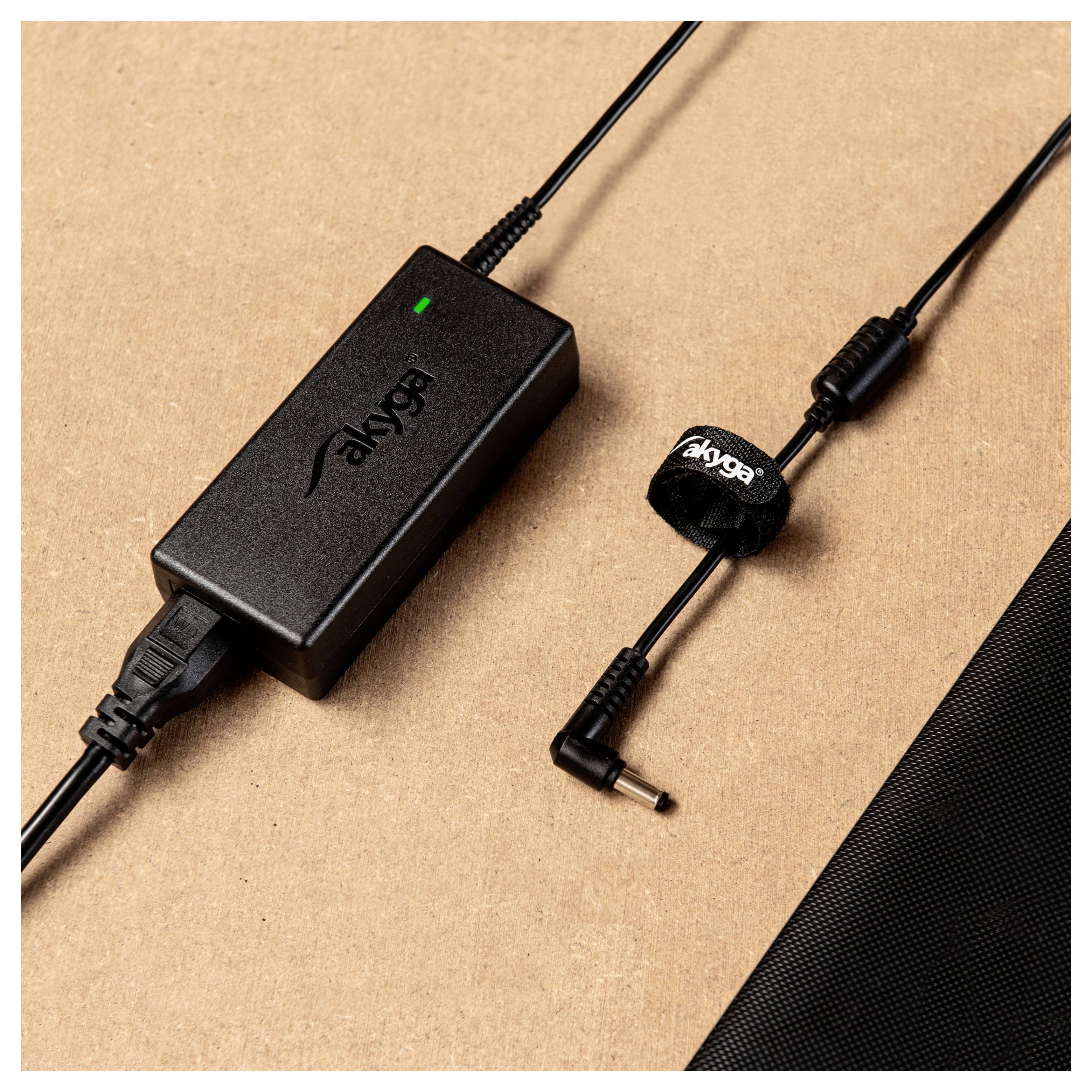 A black laptop charger with the 'akyga' logo is lying on a brown surface next to a cable with a Velcro fastener.