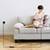 A woman is sitting on a sofa and reading a book, while her mobile phone is being charged via a charging cable plugged into a wall socket.