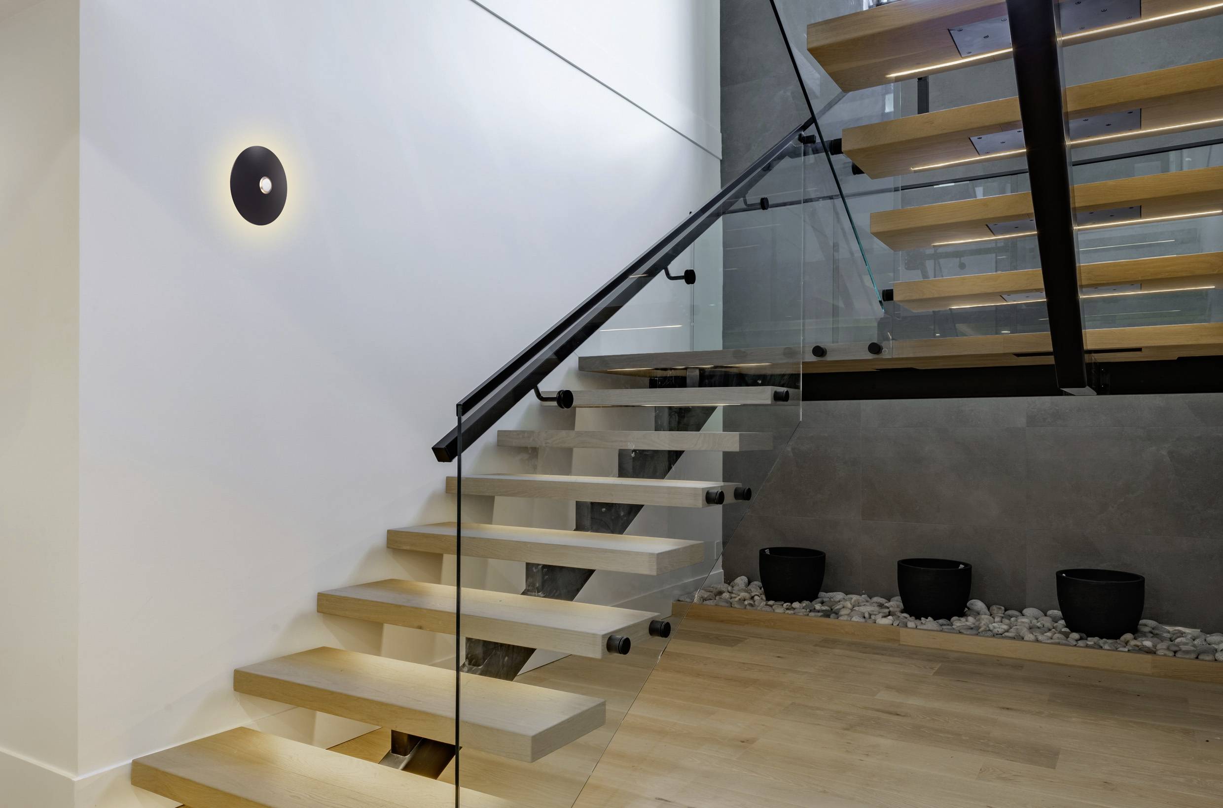 A modern open staircase with wooden steps and glass railings is shown, featuring a sleek wall light and potted plants on the landing.