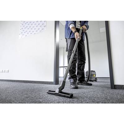 A person is hoovering a carpeted floor in an office. A table is visible in the background. The scene depicts cleanliness and order.
