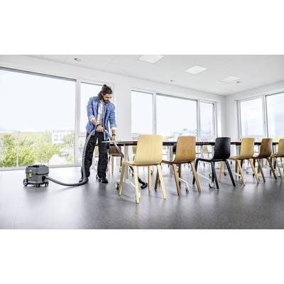 A person is hoovering the floor in a bright conference room with large windows. Tables and chairs can be seen in the background.