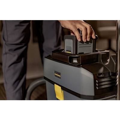 A person is inserting a battery into an industrial vacuum cleaner. The vacuum is grey with a yellow element and has a robust construction.