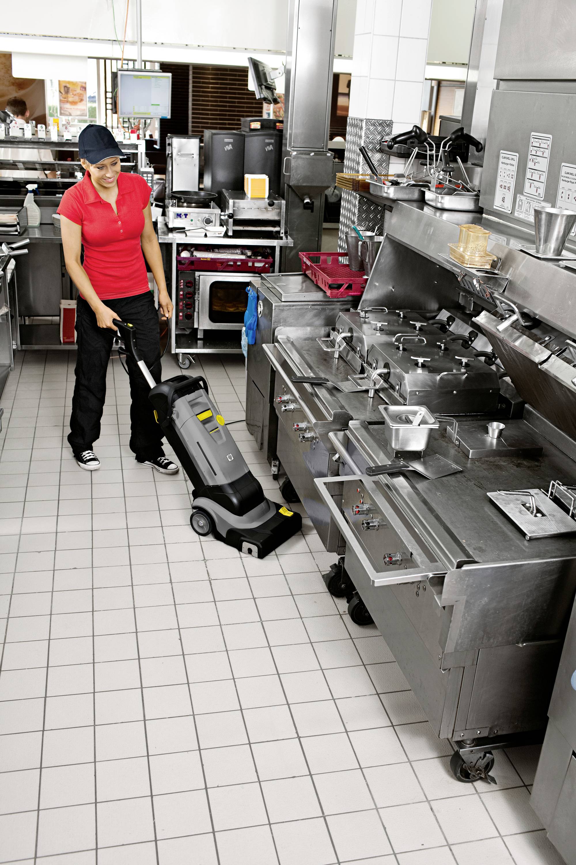 A person is cleaning the kitchen floor with a machine. The kitchen contains large stainless steel appliances and looks tidy.