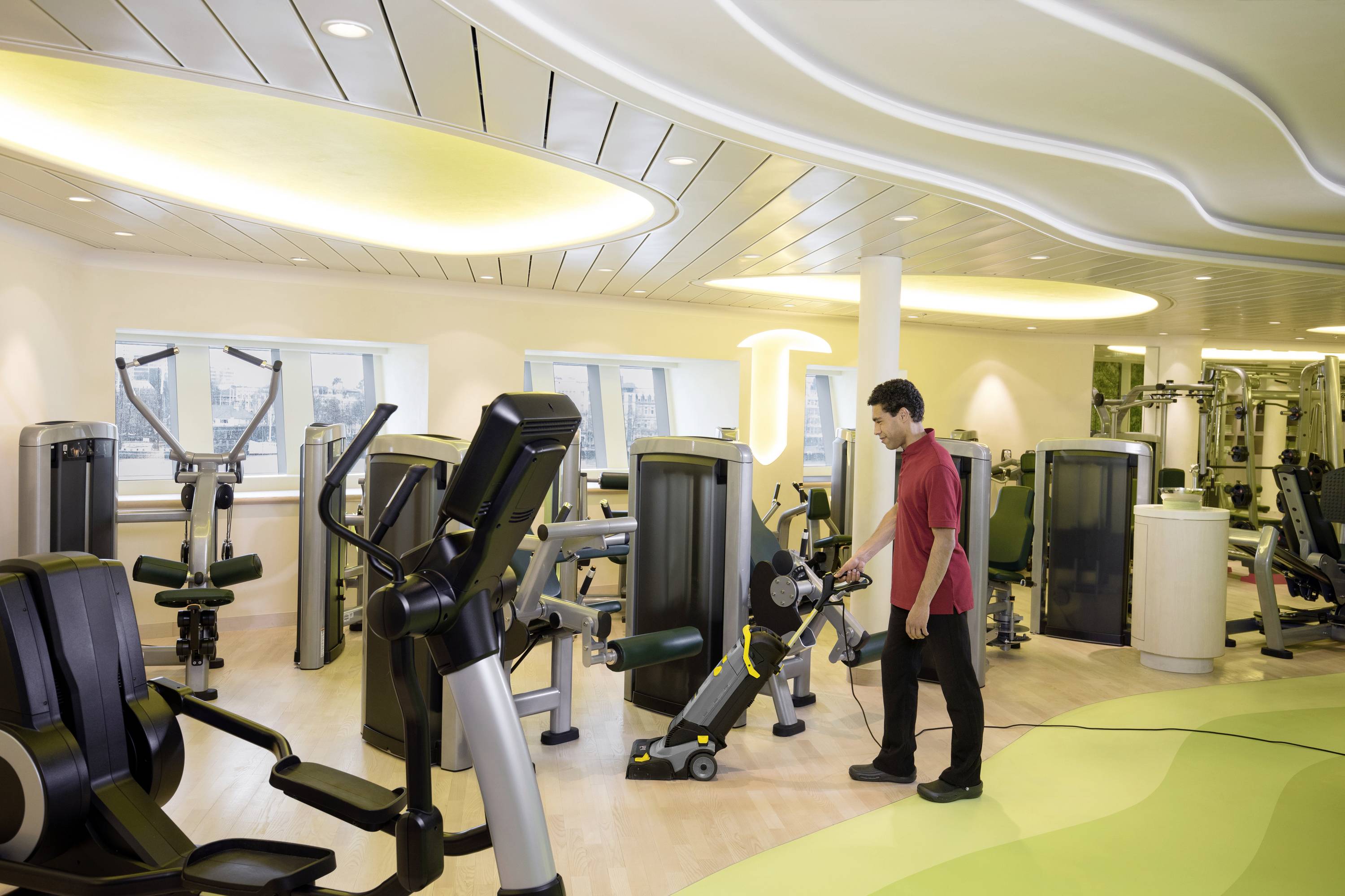 A man in a red shirt is cleaning the floor in a bright gym with various exercise equipment.