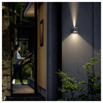 Nighttime exterior of a modern house lit by a wall sconce, with a woman using a treadmill or exercise machine on the porch in the background.
