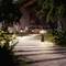 Garden path with lanterns that illuminate in the evening. In the background, a person is sitting at a wooden table in front of a house.